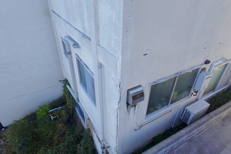 <p>長野県飯田市にて、ドローンを活用した無料建物診断を実施しました。<br>

今回は、地上からでは確認しにくい屋根や高所の外壁を中心に、建物全体の劣化状況を確認しました。<br>

屋根には赤系の金属屋根が使用されており、ドローンで上空から確認したところ、塗膜の色あせや退色、表面のムラなど経年劣化が広範囲に見られました。<br>

外壁では、全体的な汚れや黒ずみに加え、建物の角部分に縦方向へ大きく伸びるひび割れが確認できました。一部では表面が剥離して下地が露出している箇所もあり、今後さらに劣化が進行すると雨水が浸入する原因となる可能性があります。<br>

また、窓周辺や外壁の取り合い部分にも汚れや劣化が見られました。こうした高所のひび割れや外壁の傷みは地上からでは把握しにくいため、ドローンによる確認が有効です。<br>

株式会社横井では、ドローン無料調査・横井豊文教師建物診断により、屋根・外壁・付帯部まで現在の状態を確認しています。</p><p><br></p>